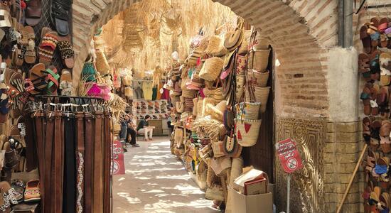 Ein belebter Souk in Marrakesch zeigt bunte Waren wie Sandalen und Körbe unter einem schattigen Bogengang.