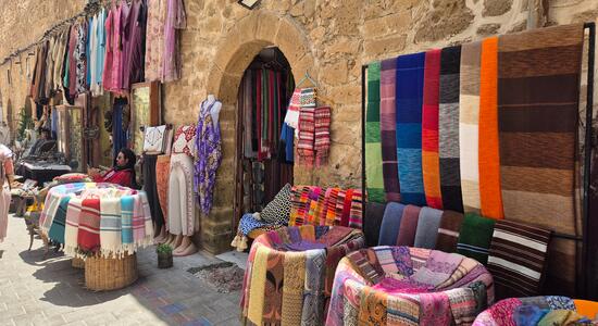 Bunte Tücher hängen und liegen auf Ständen entlang einer antiken Steinmauer in Essaouira, während Menschen vorbeigehen.